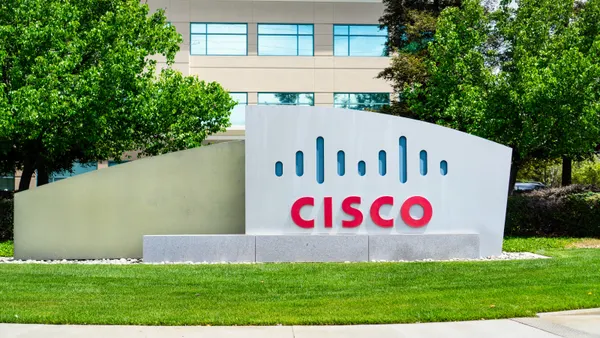 A large sign says "Cisco" in red letters. The sign sits on a grassy lawn in front of a building with many windows.