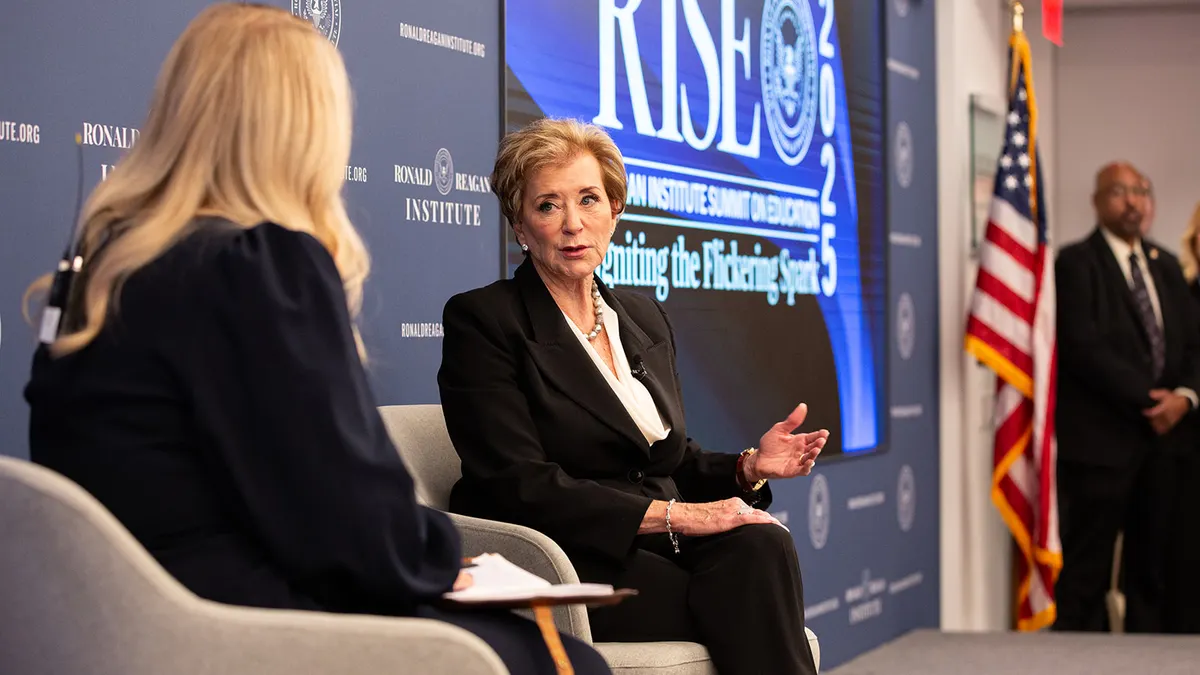 Two people are seated on a stage. One person's back is to the camera and one person is facing toward the camera. A backdrop and screen say "RISE"