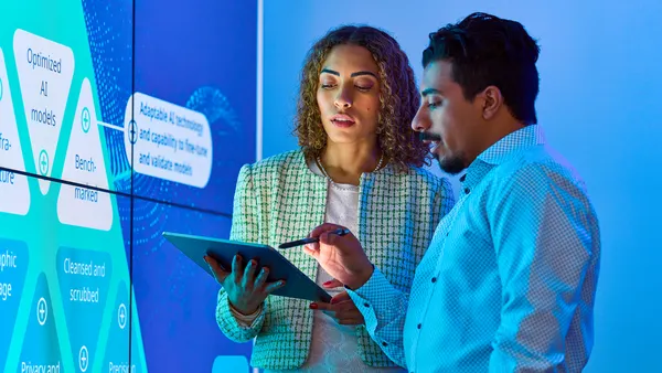 Two people in a blue-lit room discuss data on a tablet. A large digital display with AI-related keywords is in the background. The setting is futuristic and focused.