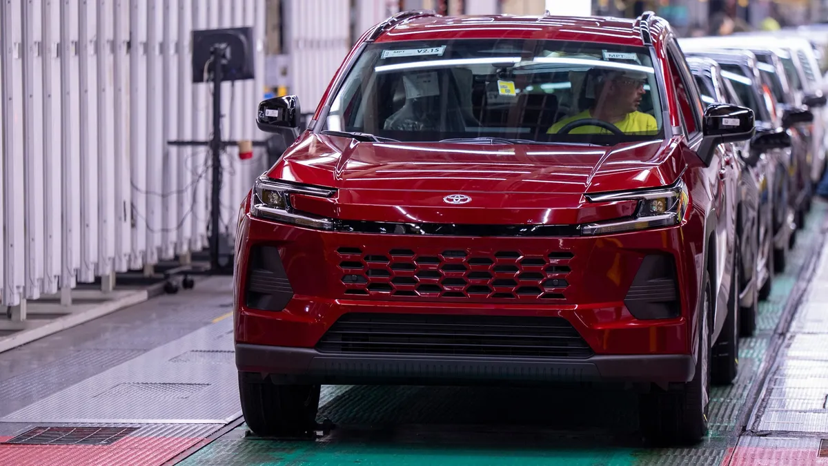A dark cherry red SUV on a factory assembly line, with a worker wearing a bright yellow shirt and protective eyewear behind the wheel.