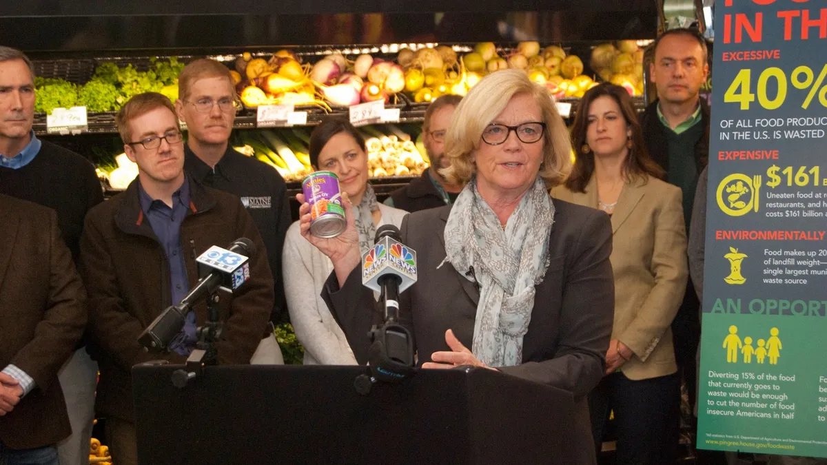 Pingree stands in front of a podium holding a canned food item in front of the produce section of a store.