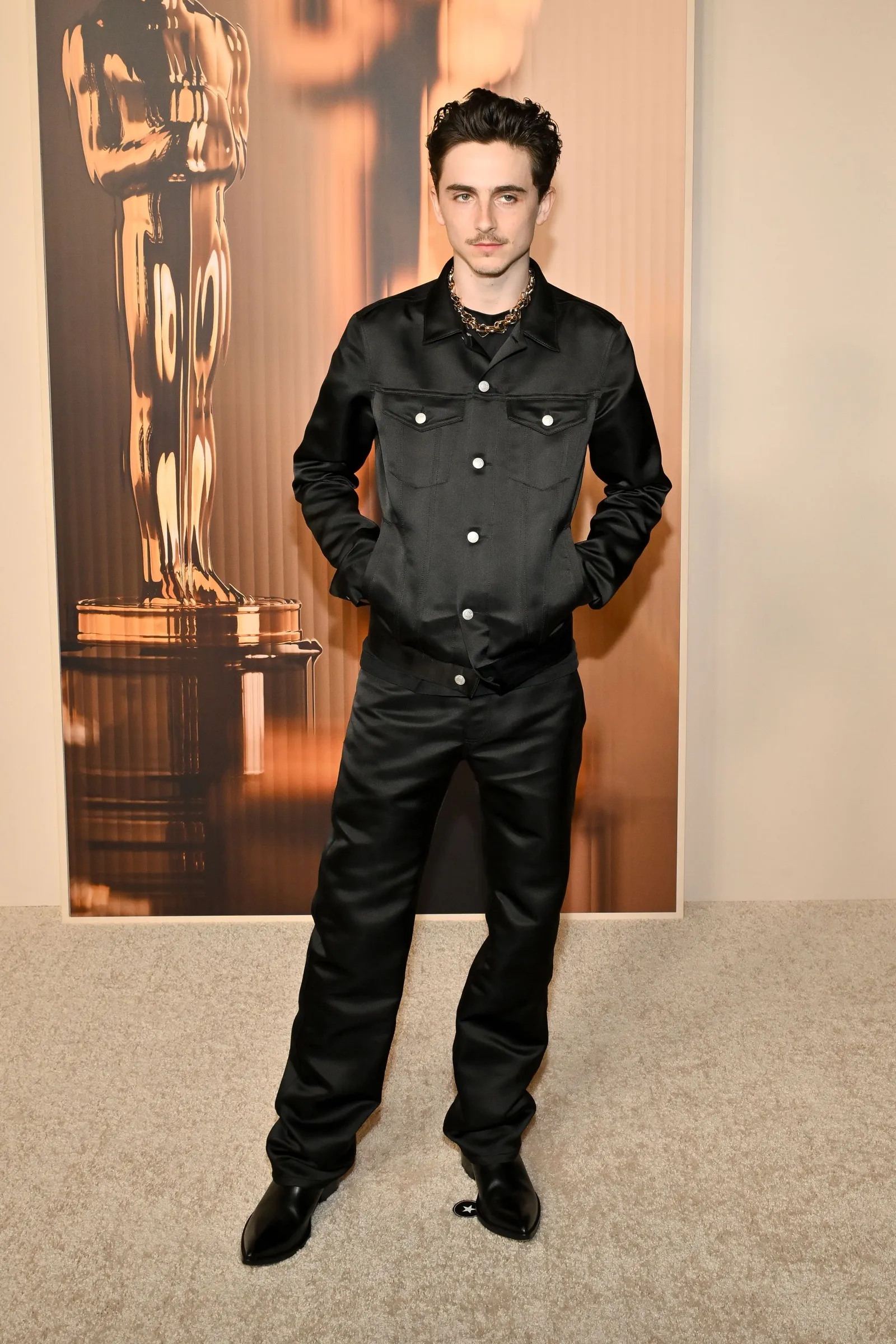 A person in a black satin suit with a cropped jacket stands in front of a gold backdrop.