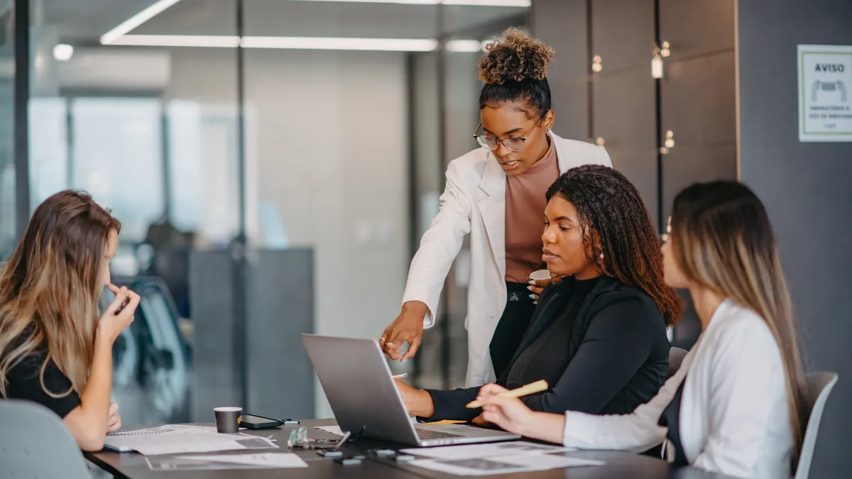 A diverse group of women gather around a computer at a table in a meeting room.