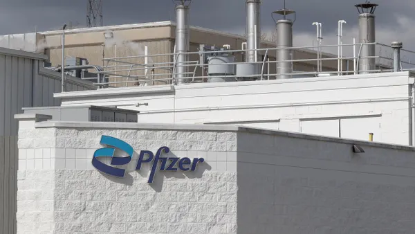 A "Pzifer" sign is mounted to a white-bricked building wall; behind the wall is the infrastructure of an industrial plant. 