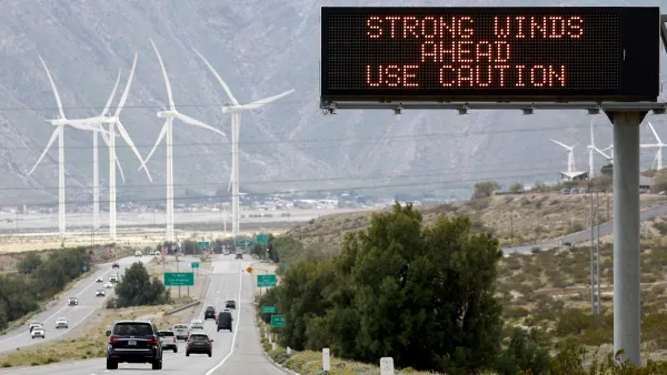 Cars driving along highway with sign in foreground that says "Strong Winds Ahead, Use Caution" and wind Turbines in the background beyond the highway.