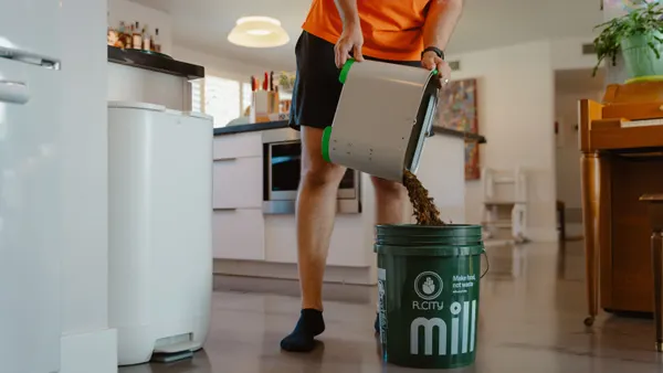 A person standing in a kitchen pours dehydrated food grounds into a 5-gallon bucket labeled with "mill" and "r.city"