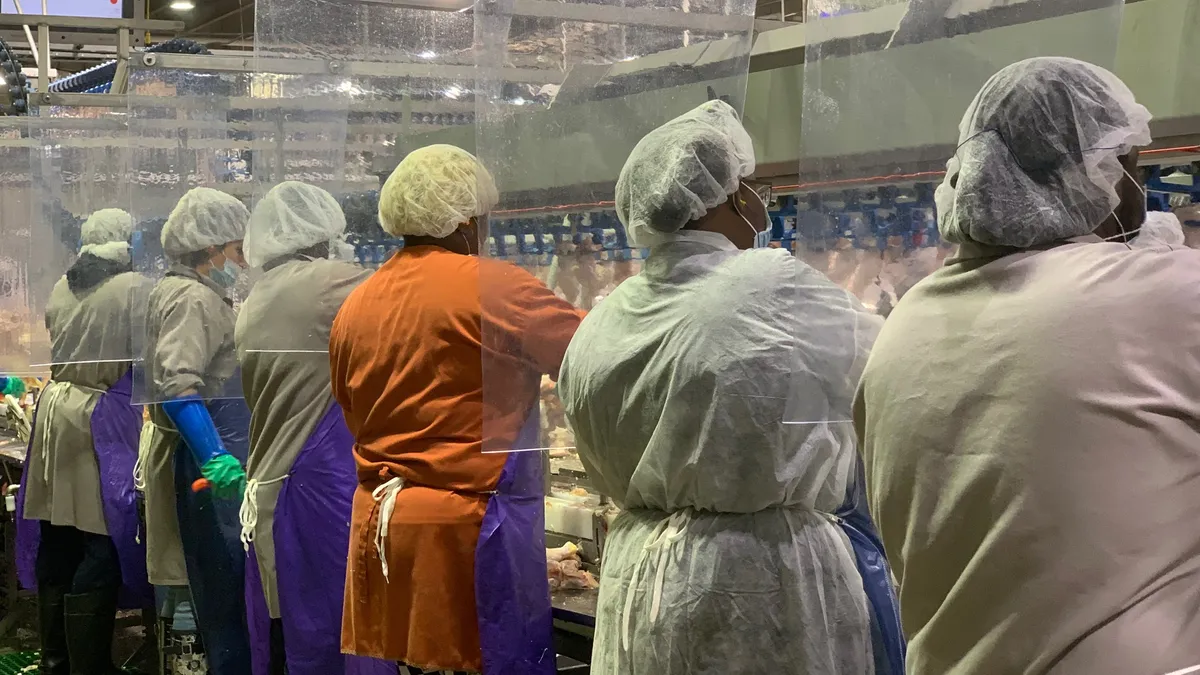 Several workers in protective gear work on a meat production line with plastic dividers between them.