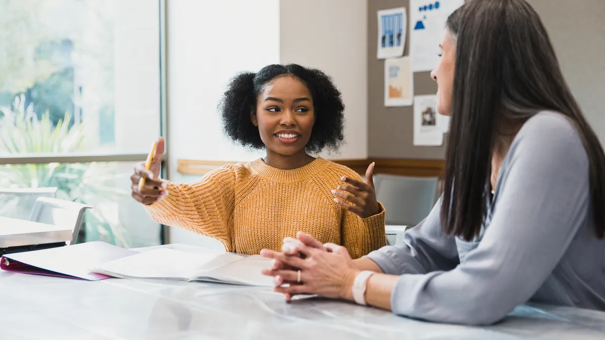 A high school student talks with her teacher sitting together at a desk in a classroom.