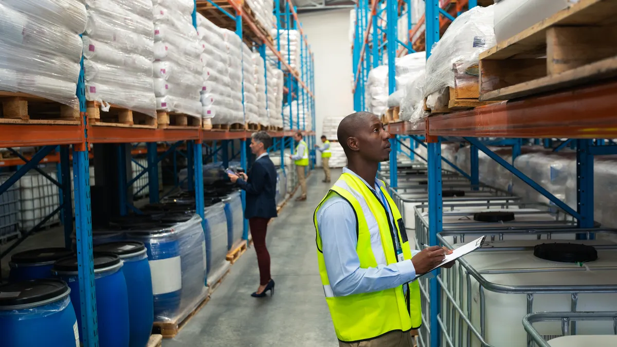 Side view of warehouse staff checking stocks in a warehouse aisle. They are holding clipboards and writing on them.