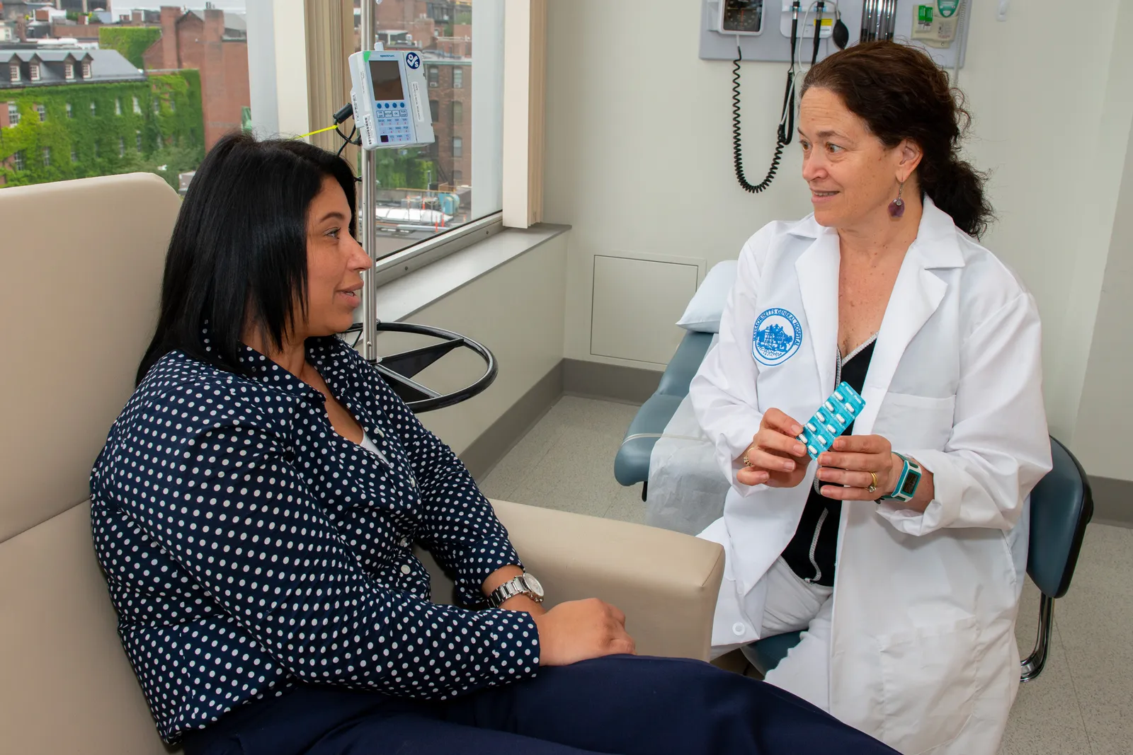 A person in a medical coat speaks to another person while seated.