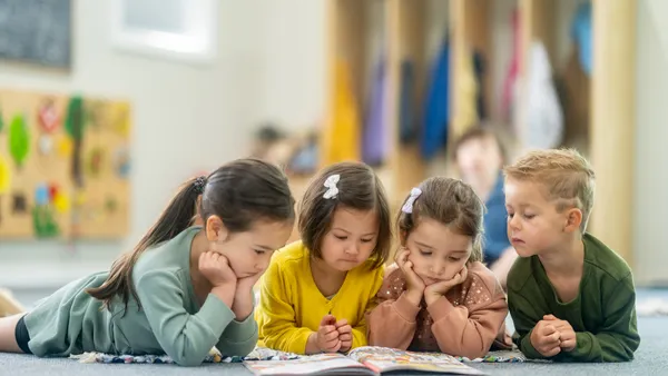 Four young children lay on their stomachs on the floor of a classroom looking at a book.