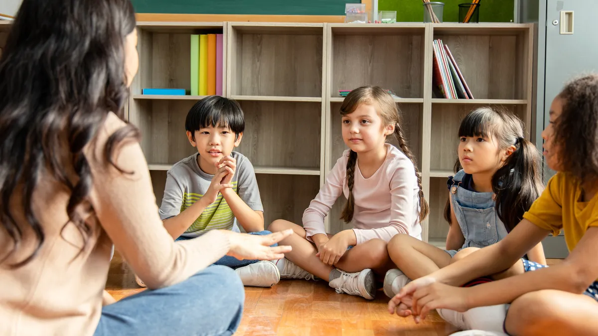 An adult sits on the floor of a classroom with their back to the camera. In front of the adult are four early elementary children who are watching the adult.