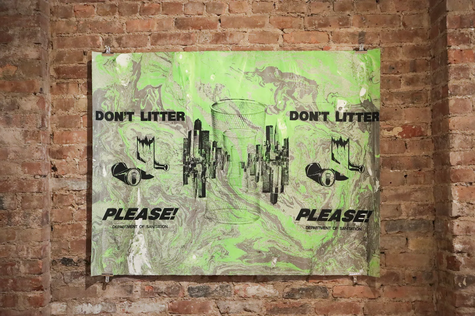 A print using repeated imagery of a waste bin, the New York City skyline, and the words &quot;Don&#x27;t Litter Please!&quot; and &quot;Department of Sanitation&quot; against a marbled background hangs on a brick wall.