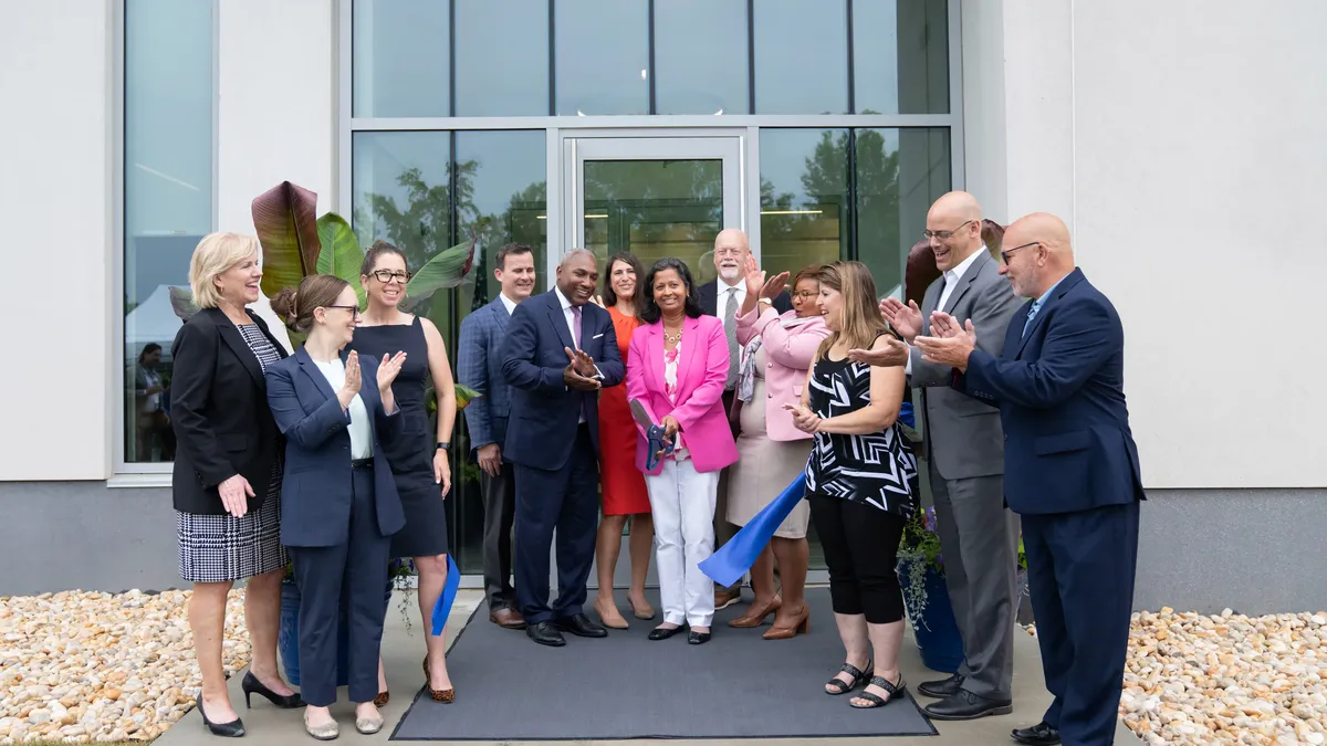 12 people in front of a white building with glass doors clapping after one person in a pink blazer cuts a blue ribbon.