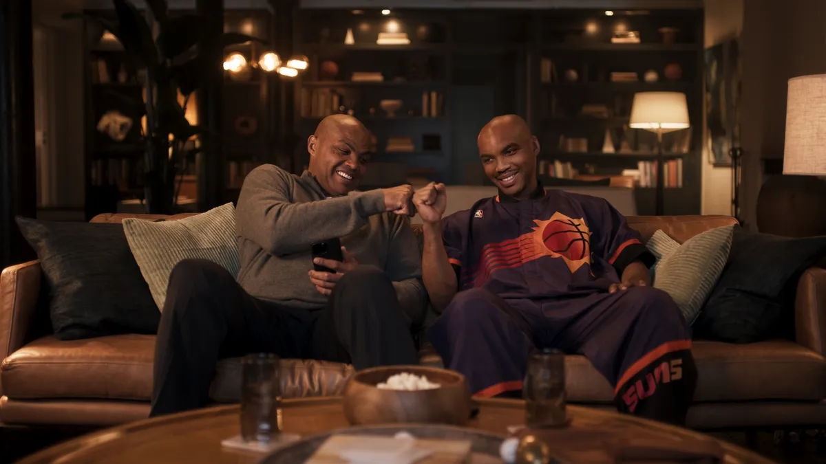 Charles Barkley sits on a couch with his young self, created by deepfake technology.