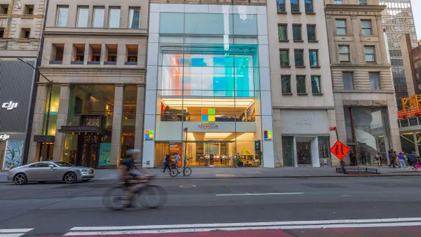 A wide view of the Microsoft Store on Fifth Avenue in New York City, featuring a modern glass façade with the colorful Microsoft logo, cyclists and pedestrians on the street in front.