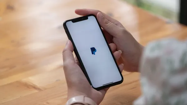 A person holds a phone at a table; the Paypal logo is on the screen.