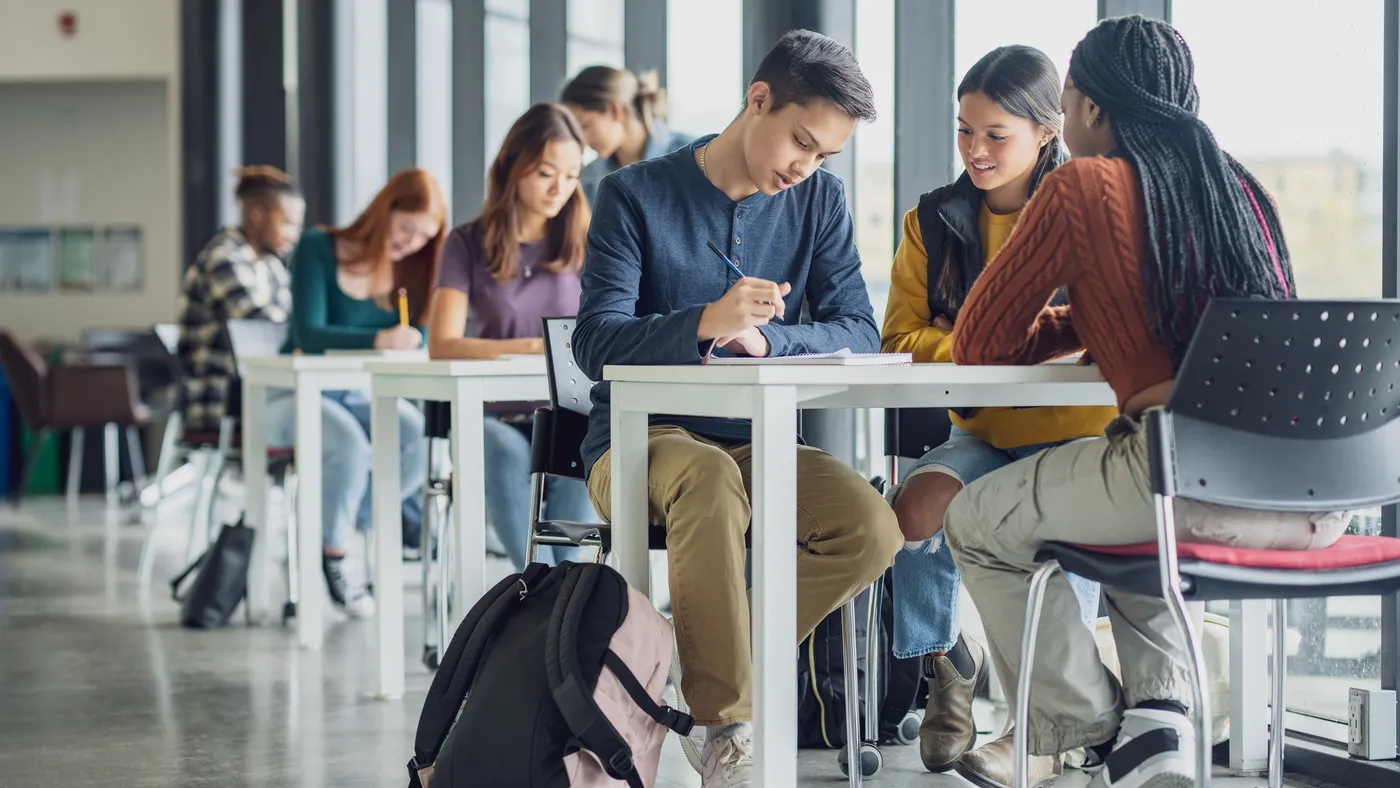 Students are sitting at several tables working in groups. There are laptops and notebooks on the tables and the tables are pushed to a wall of windows.