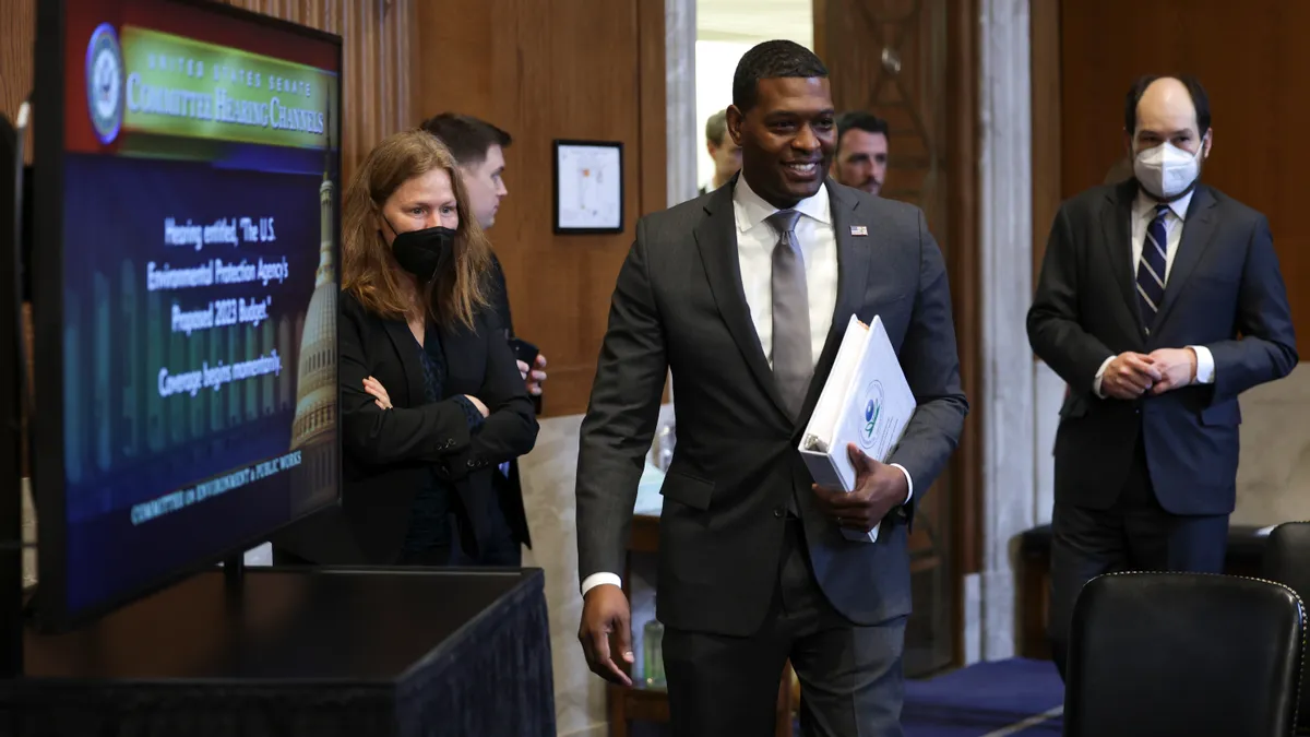 Man in suit, holding a binder, walks with a crowd of people into a room for Congressional hearing