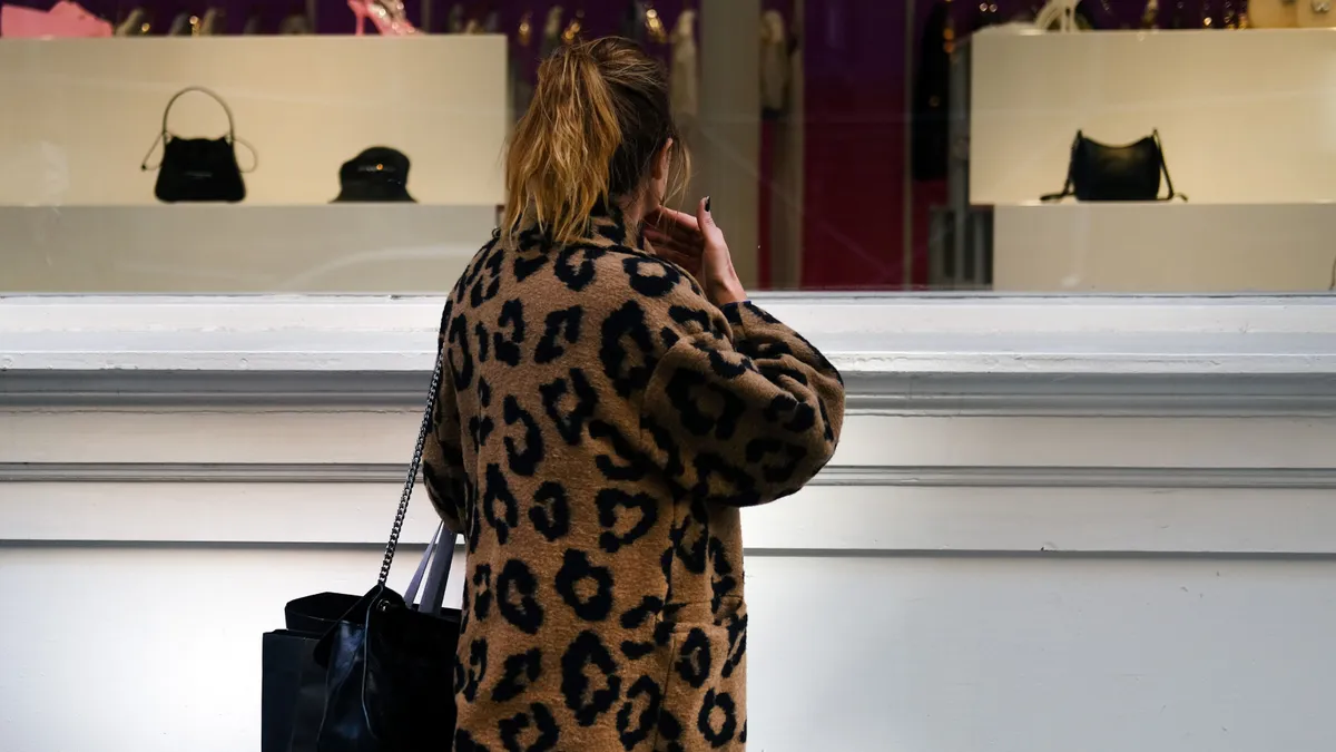 A person in a leopard print coat looks at handbags in a shop window.