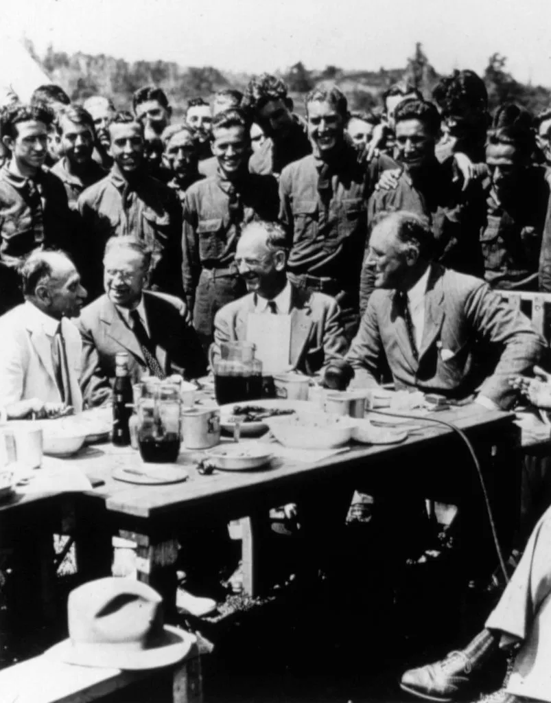 President Franklin Delano Roosevelt sits among a group of people in a Civilian Conservation Corps camp