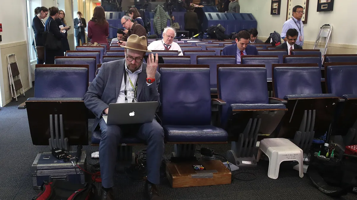 People sit in rows of chairs and mill about them. One person in the foreground holds a phone up to his ear while typing on a computer.