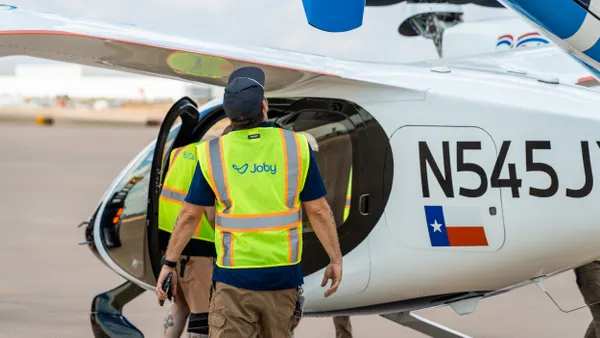 A person wearing a visibility vest walks next to a small white and blue aircraft.