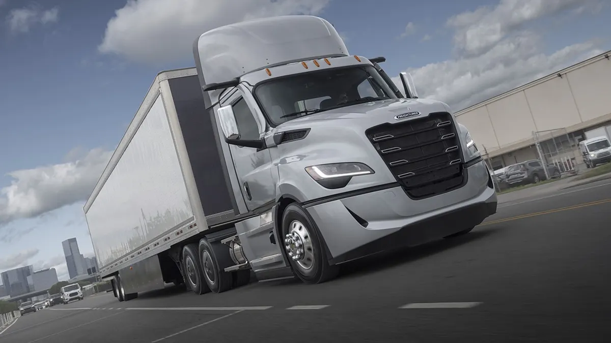 A Freightliner Cascadia's fifth generation tractor-trailer on a highway with city buildings and traffic in the background.