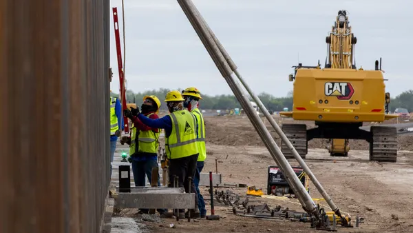 Three construction workers in bright helmets and vests stand next to a tall wall, with construction equipment in the background.