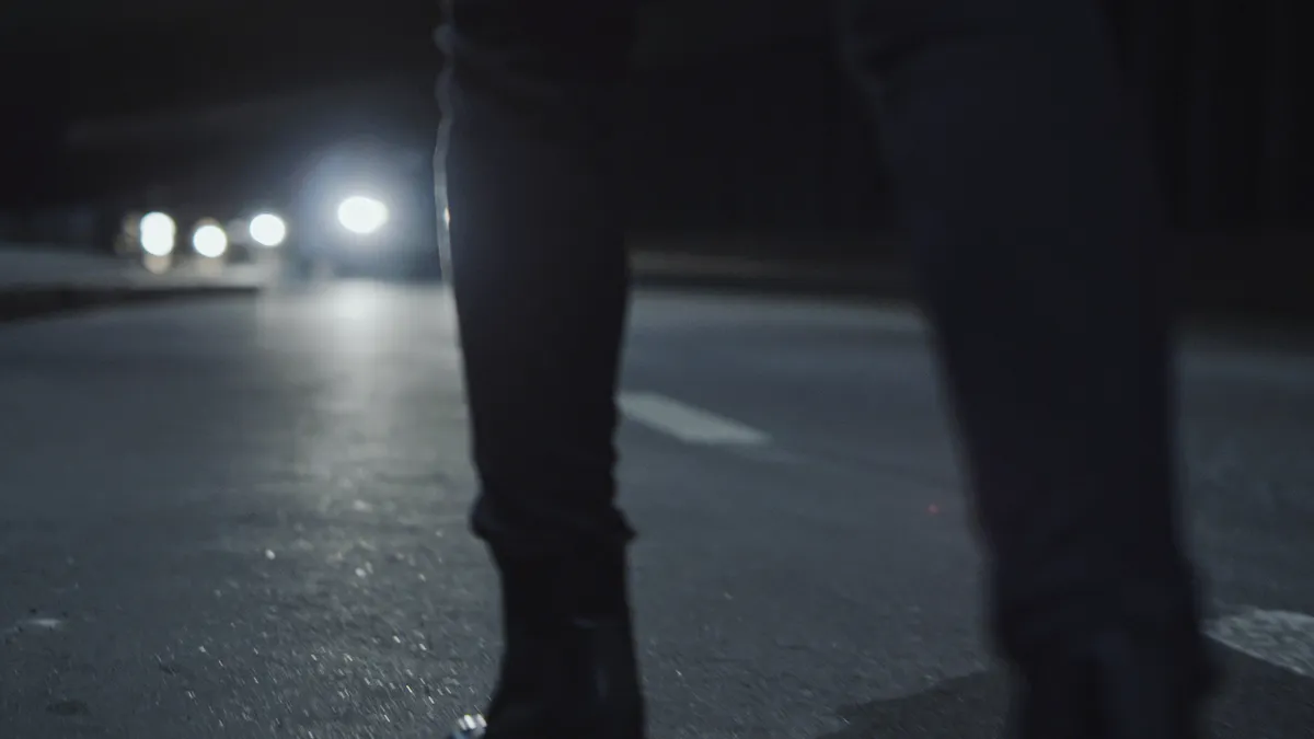 A person seen from the legs down is walking on a dark road at night with headlights approaching in the distance.