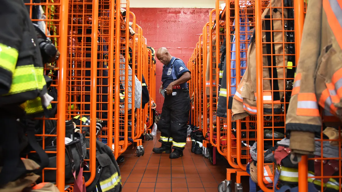 A person putting on firefighter trousers in the center, with rows of firefighter protective gear hanged on the left and right side.