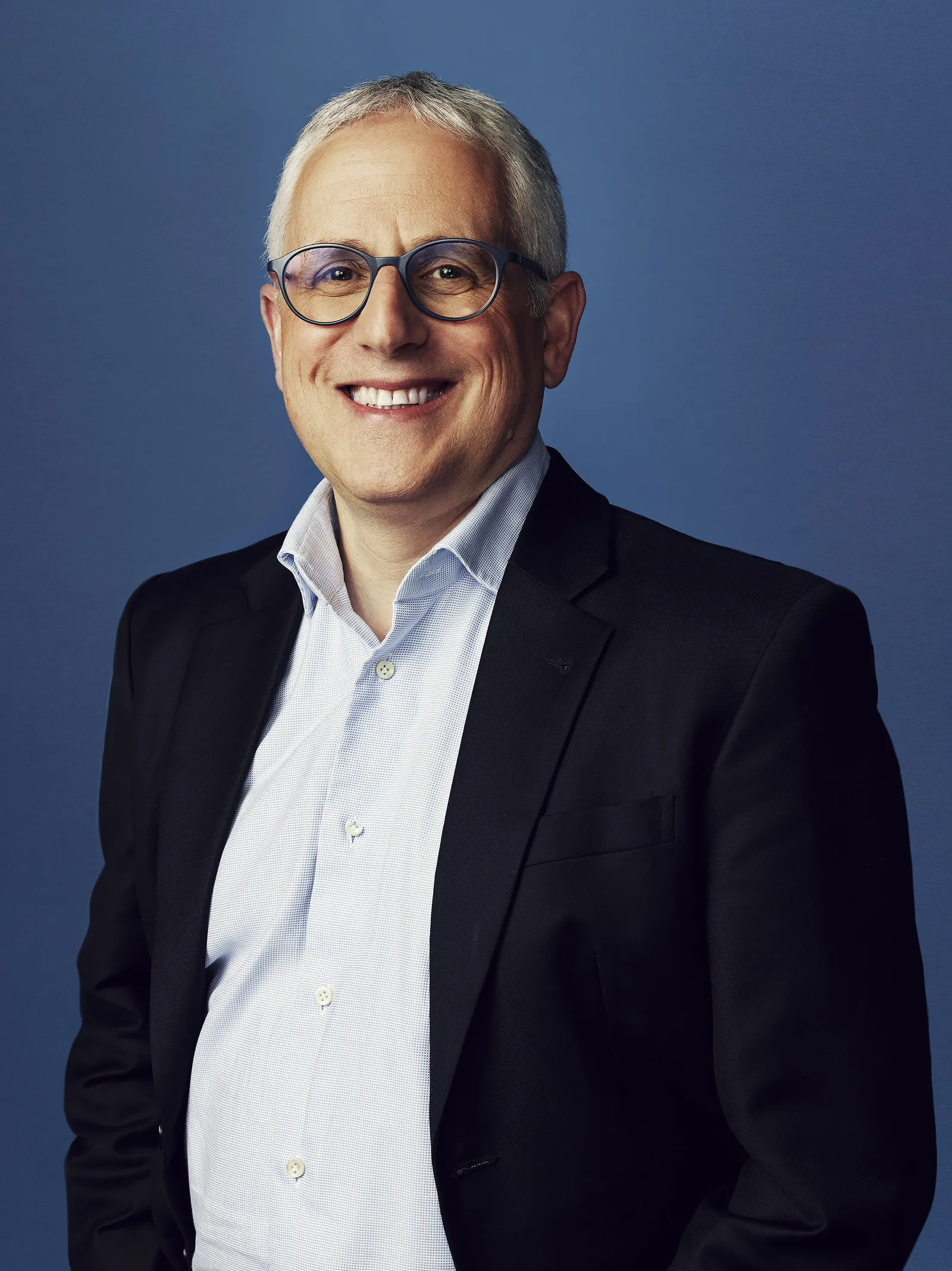 A man in a blazer and button-down shirt standing against a blue background