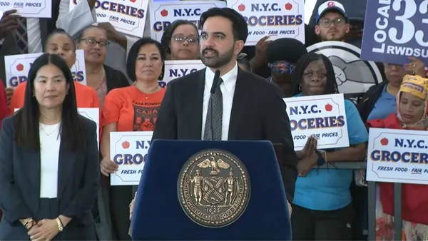 New York City Mayor Zohran Mamdani speaks while standing at a lecturn in front of people holding signs that say "N.Y.C. Groceries. Fresh Food. Fair Prices."