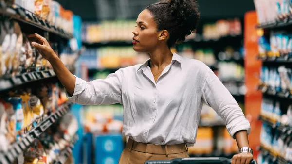 A thoughtful woman shops in a supermarket, browsing through products on the shelves. She seems to be making a careful decision about her purchase.