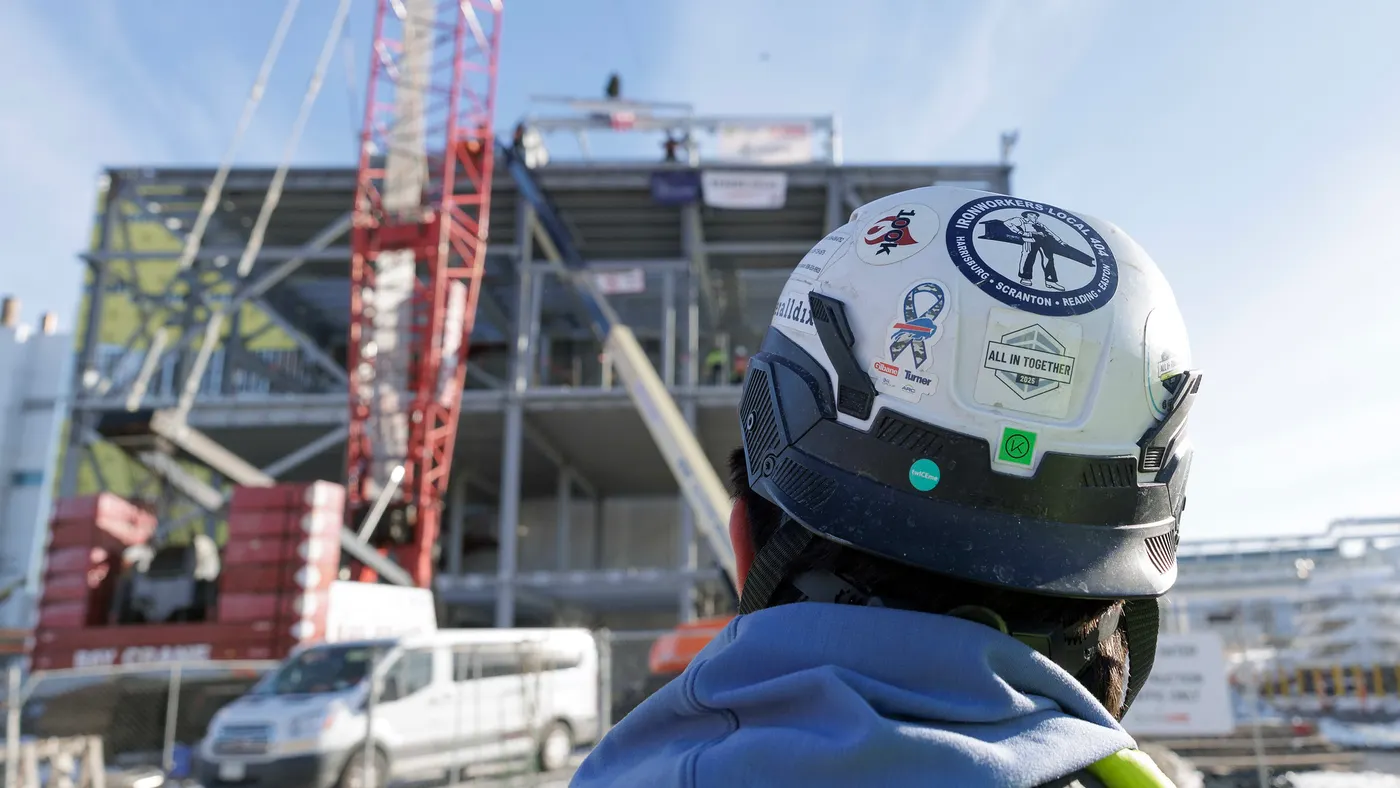 A person with a white hard hat looks up at the steel skeleton of the building in a blurry foreground.