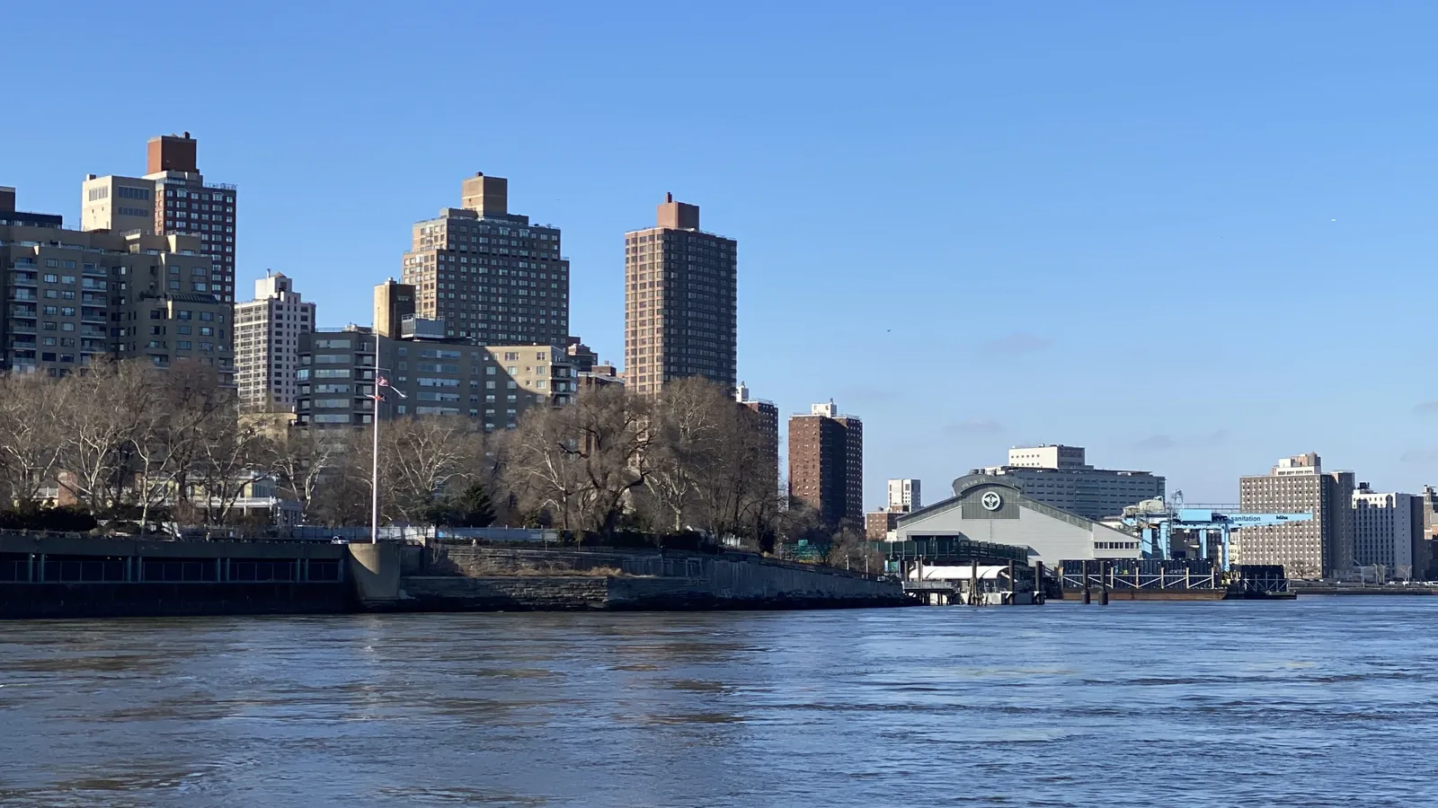 A large industrial building wih a prominent DSNY logo sits on the shore of a river next to taller residential and commercial buildings