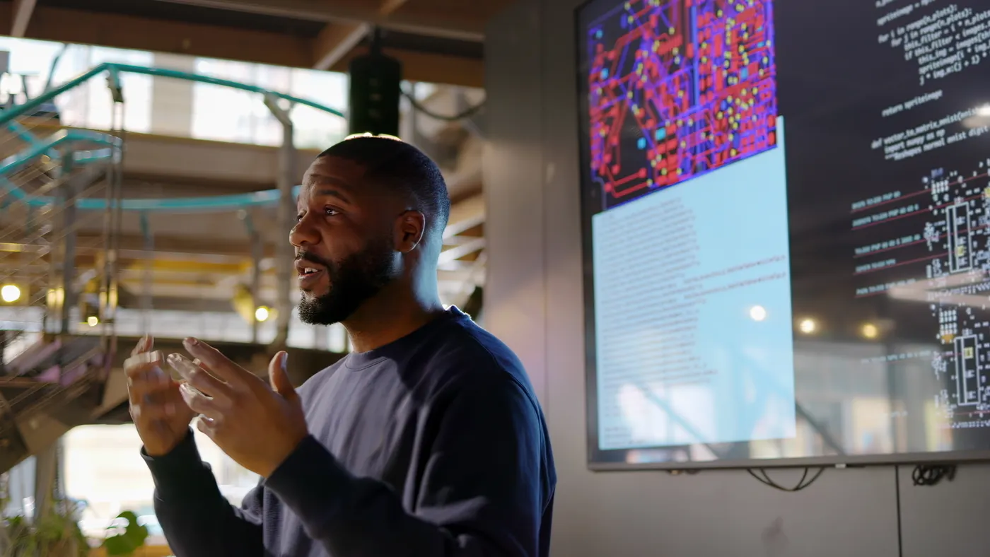 A Black man stands at a screen, teaching a seminar on Python coding.