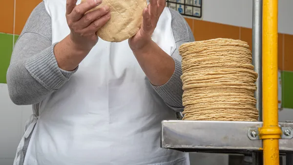 A person wearing a white apron and handling yellow dough with a stack of corn tortillas next them.