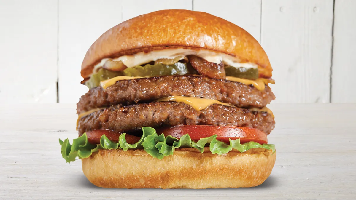 A double-decker plant-based burger with lettuce and tomato, American cheese, pickles and other toppings sitting on a white wood-grain surface with white wood paneling in the background.