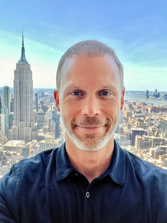 A headshot of a man in a navy button-down shirt with the New York City skyline in the background