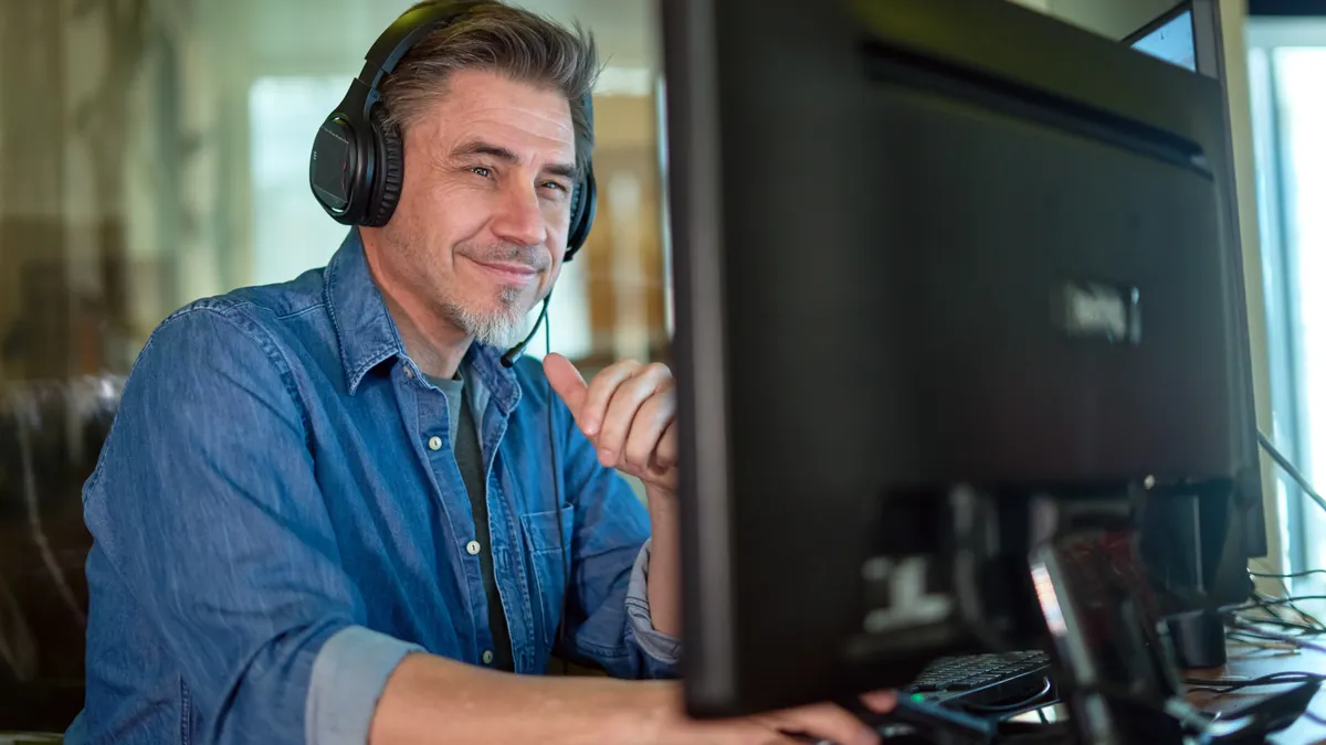 A man wearing headphones and a denim shirt sits at a desk, smiling while looking at a computer monitor. The scene conveys a focused, relaxed mood.