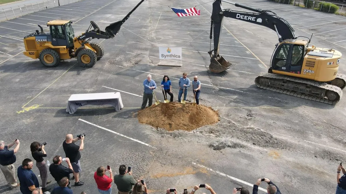 Overhead view of four people shoveling dirt in a ceremony on a dirt industrial site with photographers looking on.