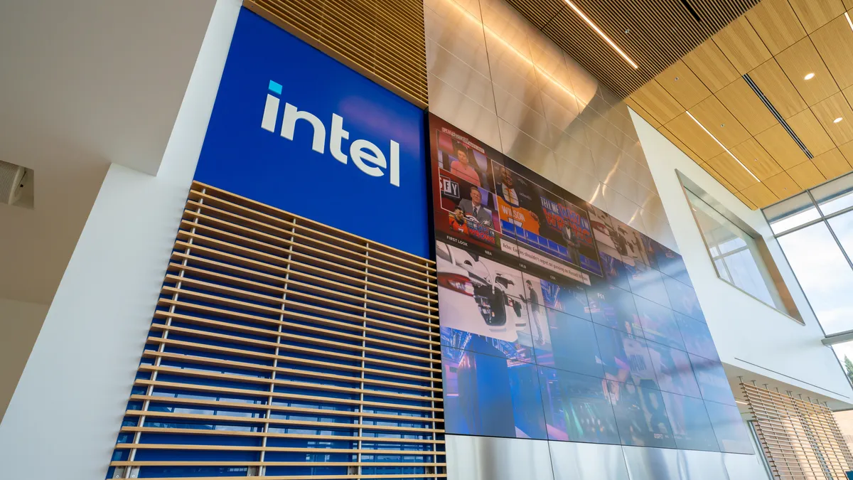 The blue Intel logo on a wall with a large TV on its left inside a building.