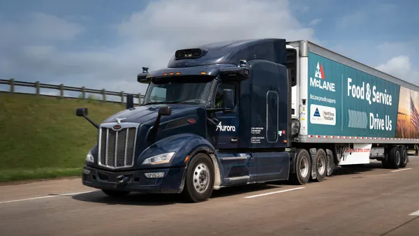 An 18-wheeler drives down a highway. The cab of the truck is dark blue and the trailer has writing on the side that says "McLane. Food & Service drive us."