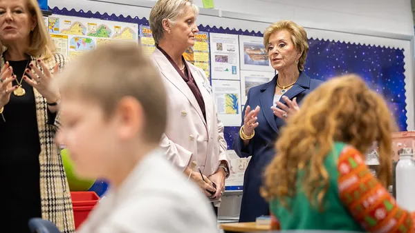 Three adults are standing in a classroom talking and listening. Children siting at desks working on assignments are seen in the foreground..