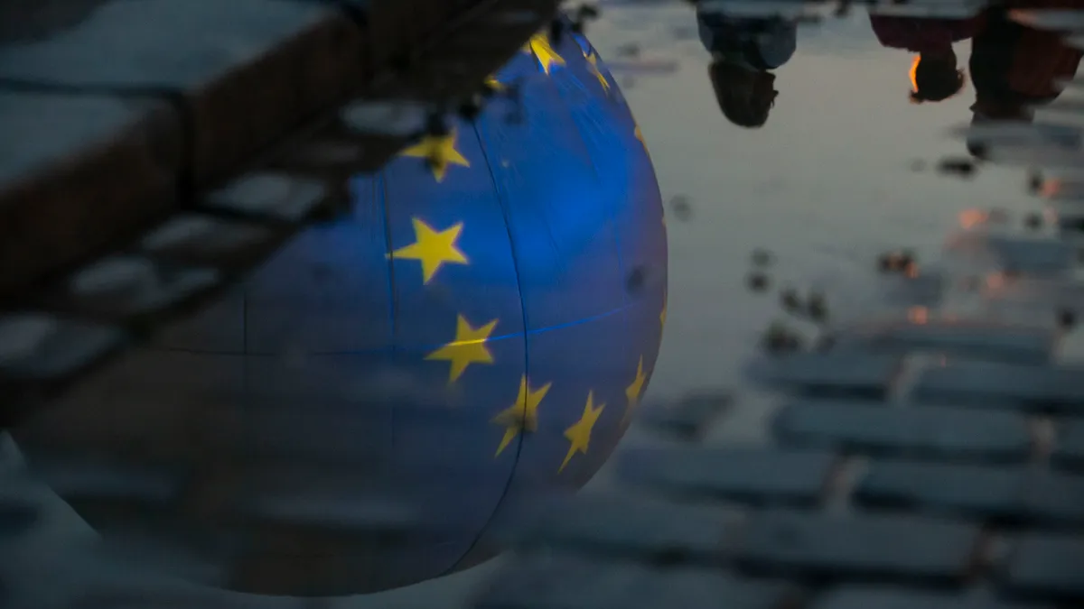 The flag of the European Union is projected on helium balloon reflected in a puddle on a cobblestone street.