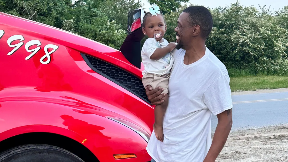 Flatbed driver Marcus Yarborough smiles while holding his 1-year-old granddaughter, Kyari, in a picture with his red truck.