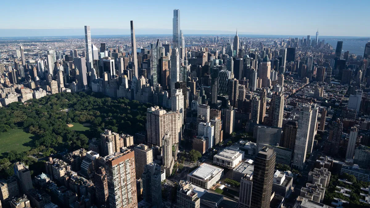 Aerial photo of Central Park and the surrounding buildings on a sunny day in New York City. The camera is angled downwards and captures a view of Central Park to the left, with the surrounding skyscra