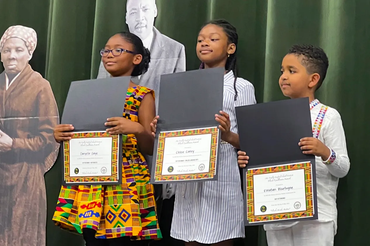 Three students stand on a stage, each holding a document in front of them. Behind them are life-size cardboard cutouts of people.