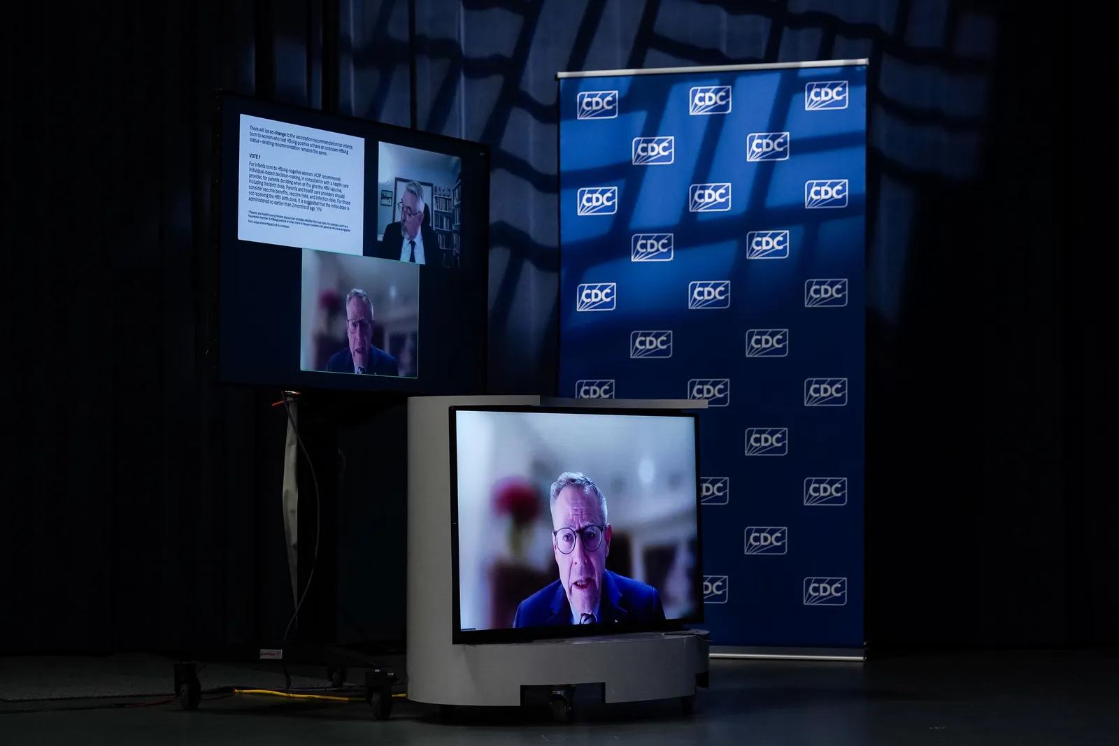 A TV monitor with a man speaking sits in front of a blue wall that has CDC printed on it multiple times with three screens in the left above the TV show the same man in one screen on the bottom, another man on a screen to the right above, and an image of text on the screen to the left.